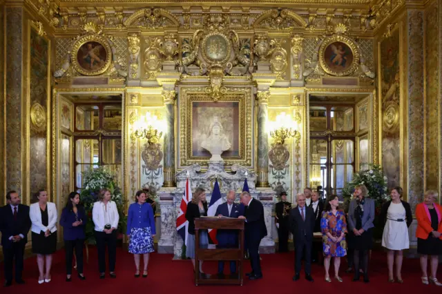 King Charles joined by others signing the visitors book in the Salle des Conference