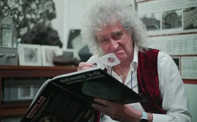 Sir Brian May holding a book titled 'Bennu 3-D' and smiling