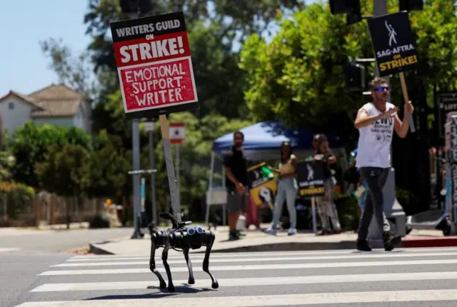 A robot dog, decorated with googly eyes, at a WGA picket line. It has a sign attached to it with the phrase: Emotional support ‘writer’.