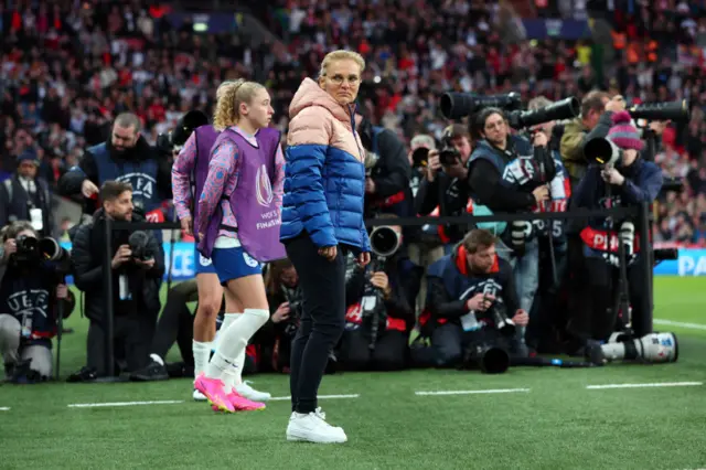 Sarina Wiegman stares at the camera as a hoard of press snap pictures behind her.