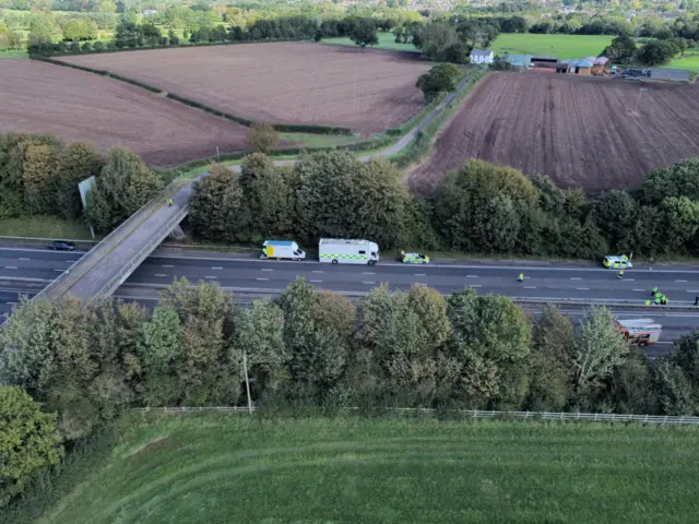 A wide shot of the motorway, with a bridge on the left and emergency vehicles parked on either side of the road, with some people in hi-vis in the road