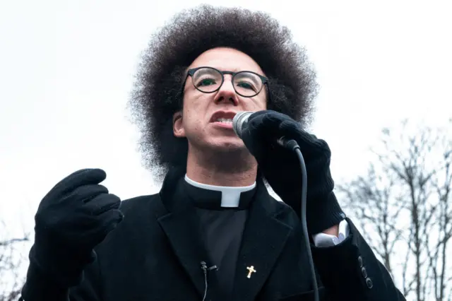 GB News presenter Calvin Robinson is seen with a microphone, wearing black gloves and a black coat