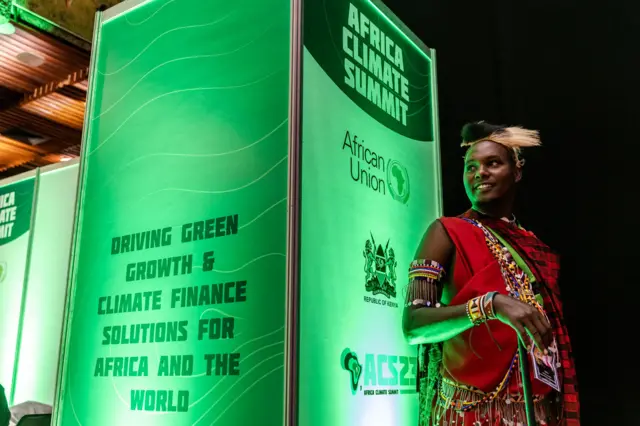 A Maasai man stands next to some Africa Climate Summit logos while waiting for the opening session to start during the Africa Climate Summit 2023 at the Kenyatta International Convention Centre (KICC) in Nairobi on September 5, 2023