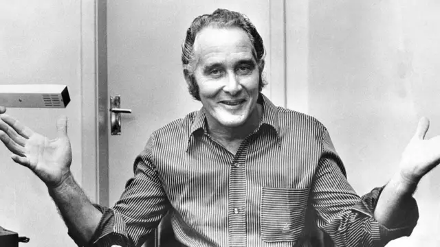 Ronnie Biggs sitting at a desk smiling and gesturing with his arms