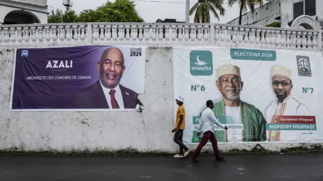 Pedestrians walk past a banner (L) of incumbent Comoros President and presidential candidate for the Convention for the Renewal of the Comoros (CRC) party, Azali Assoumani, in Moroni, on January 12, 2024.