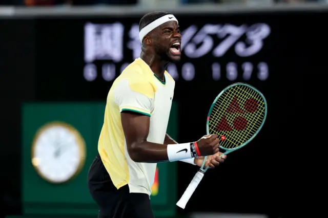 Frances Tiafoe screams and pumps his fist in celebration, wearing a white Nike shirt with yellow print on the sleeves and back.