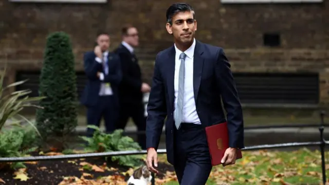 Sunak walks through a park in Westminster, carrying a red folder