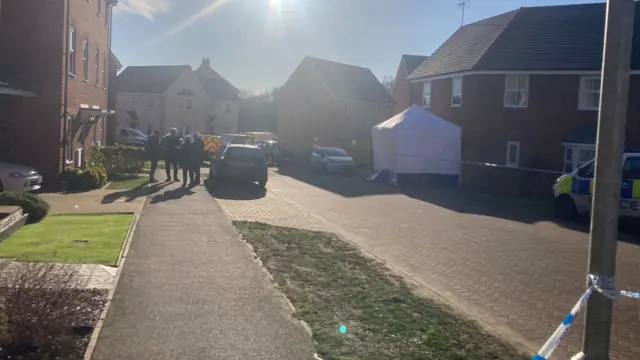 Police officers are opposite a house taped off and with a white tent outside.