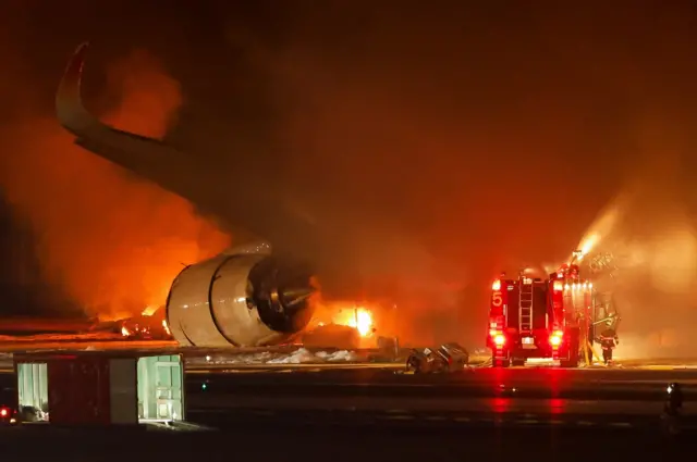 A fire truck sits behind the wing of the plane, with plumes of orange smoke filling the air