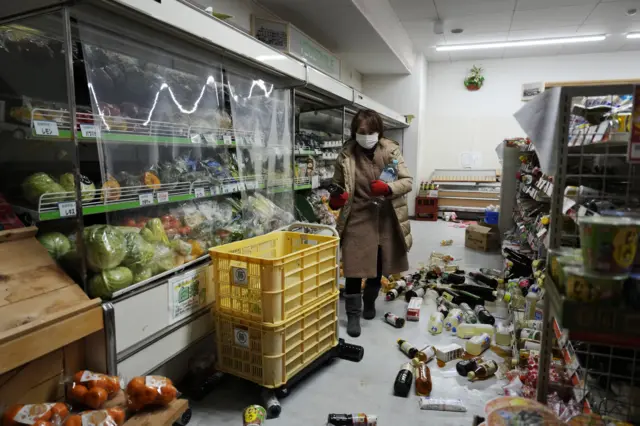 A woman walks past goods strewn about in a damaged grocery store after a strong earthquake in Togi tow