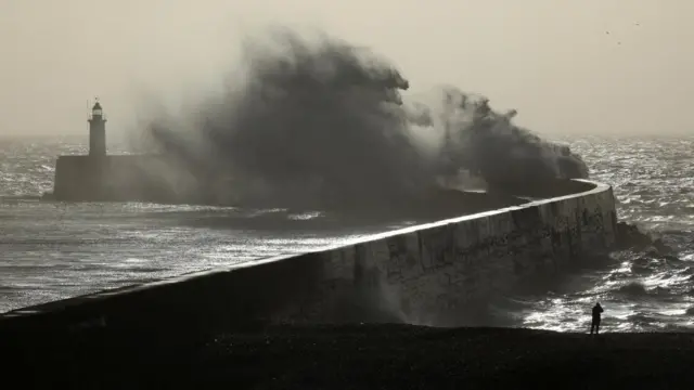 Waves crash against a sea wall with a lighthouse visible behind