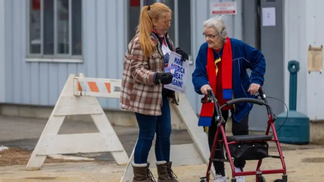 An elderly woman using a walker speaks to a woman carrying a Trump "make America great again sign" and a leaflet outside a polling station