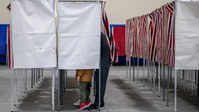 Adult and child inside a polling boot with the curtains closed. The child is on the tip of their toes.