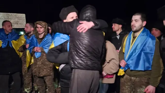 A released Ukrainian PoW hugs another man. Other released men are in military fatigues and draped in Ukranian flags.