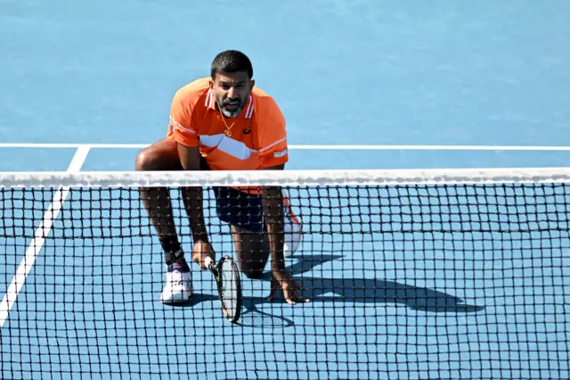 Rohan Bopanna crouches down in front of the net wearing an orange polo shirt with white patterns and black shorts