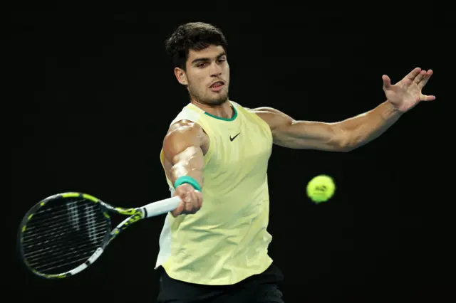 Carlos Alcaraz hits a forehand shot wearing an off-yellow Nike vest and black shorts