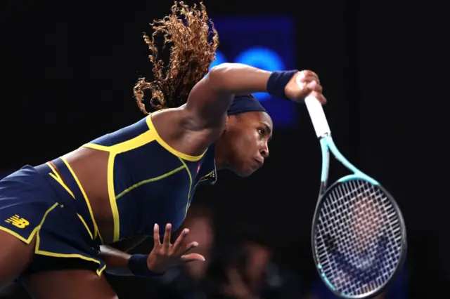 Coco Gauff serves wearing a dark blue New Balance top and skirt with yellow trim