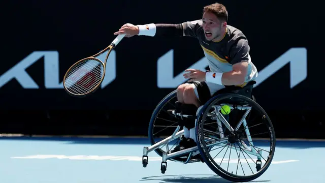 Alfie Hewett serves wearing a t-shirt with a dark green top secrion, thin yellow and black stripes and an off-white bottom section and white sweatbands with the right wheel of his wheelchair slightly off the ground