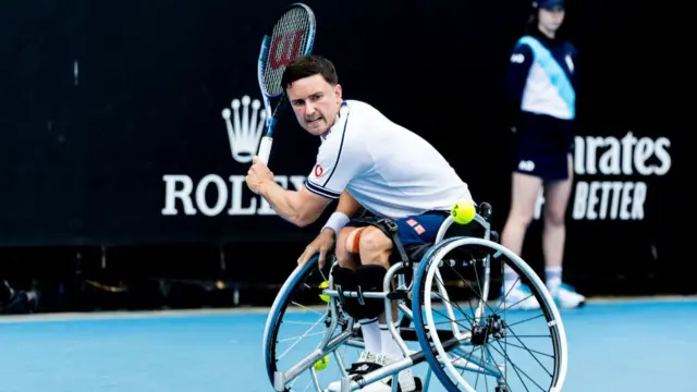 Gordon Reid plays a backhand slice shot wearing s white shirt with thin blue trim on the sleeves and navy blue shorts