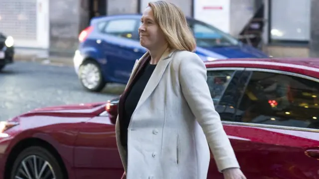 Liz Lloyd wears a beige jacket, walking briskly past a red car