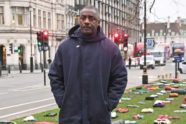 Idris Elba standing solemnly with his hands in his pockets in front of anti-knife crime campaign where 200 bundles of clothes are laid in Parliament Square