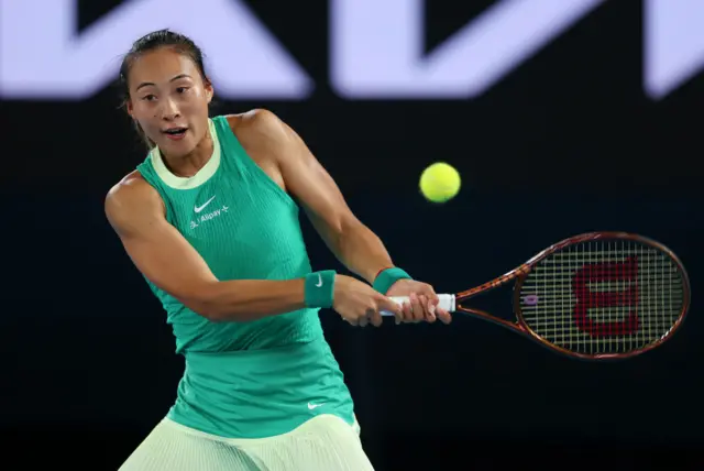 Qinwen Zheng hits a backhand shot wearing a lime green Nike vest top and cream yellow skirt with lime green Nike sweatbands