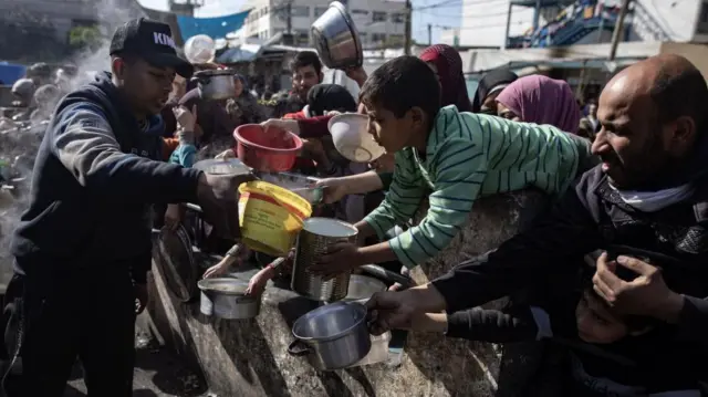 An aid worker pours food into buckets and pots held out by a crowd of men women and children