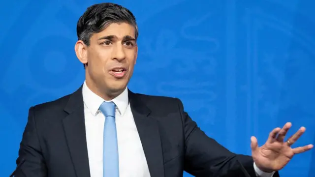 Rishi Sunak reaches out his hand in gesture as he speaks in front of a blue backdrop