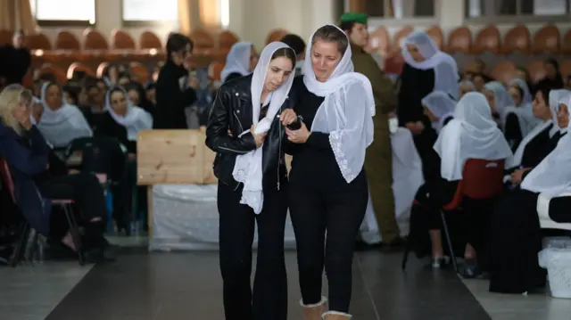 Two women wearing white headscarves support one another