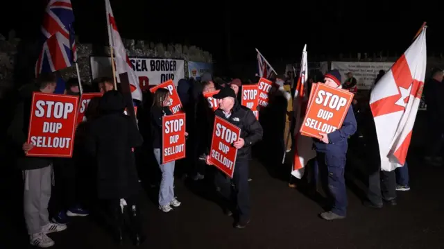 A number of protesters pictured at night, some of whom hold placards saying: "Stop DUP sellout"