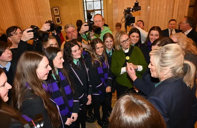 School kids huddled in a circle around Mary Lou McDonald and Michelle O'Neill amongst cameras