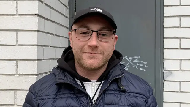 A man with glasses stands in front of a grey door in a white brick wall. He's wearing a dark blue puffer jacket over a white tracksuit top, and a North Face baseball cap. He's smiling slightly.