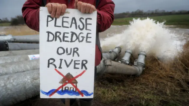 A protester holds up a sign reading "please dredge our river"