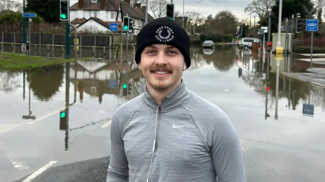 Harry Allitt out for a run. The 22-year-old wears a grey running top and black beanie hat. Behind him, the road is covered in water.