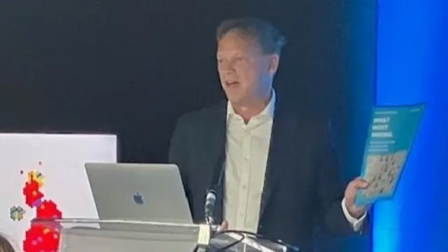 Grant Shapps standing at a lectern, in front of a laptop. He is speaking and holding a report with the title "What Went Wrong"