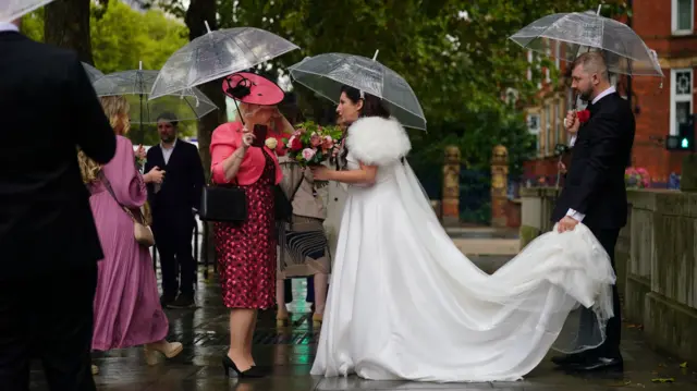 A bride in a white wedding gown stands with onlookers in the rain under an umbrella