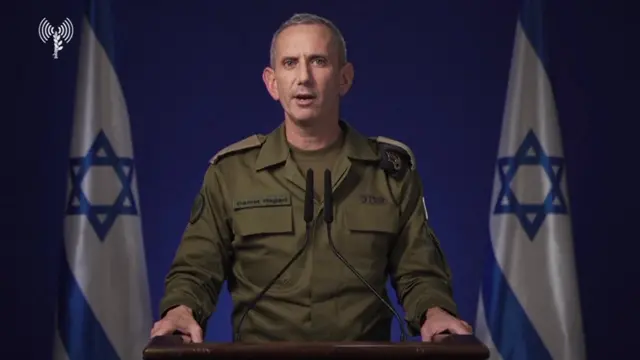 Daniel Hagari speaking into a microphone in front of two Israel flags