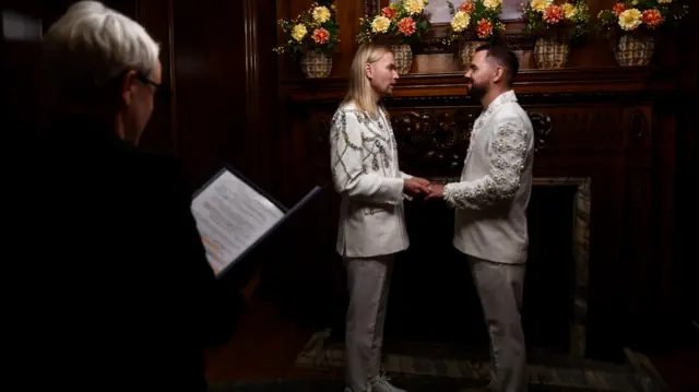 A couple wearing white outfits hold hands and face each other as they exchange vows.