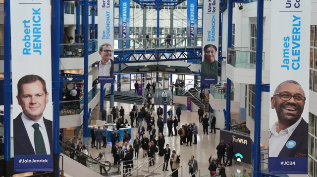 A picture of the hall where the conference is being hosted. Four banners can be seen. They are of each politician in the running for tory leadership. They contain the name and a headshot of each MP.