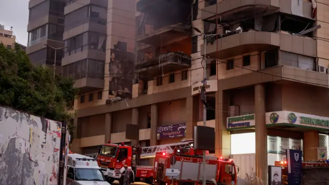 Firefighter trucks at the bottom of a damaged block of flats with holes and smoke