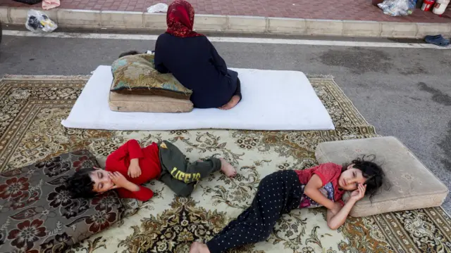 A small boy and girl lie on a carpet in a carpark, with their mother sitting with her back to them.