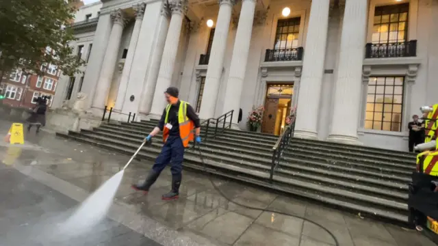 A cleaner in a hi-vis jacket power washes outside the steps at the town hall