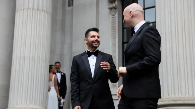 A couple wearing black suits smile at each other