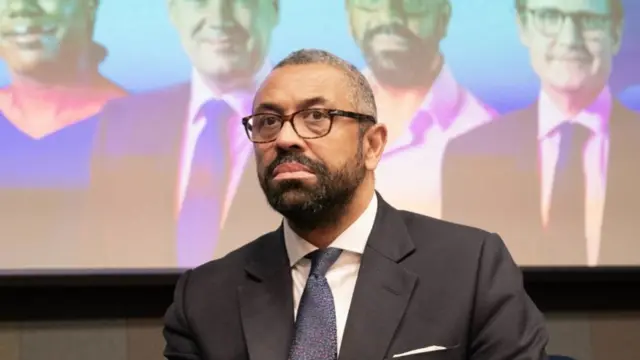 Close up of James Cleverly sitting on stage wearing black suit, white shirt and dark blue patterned tie