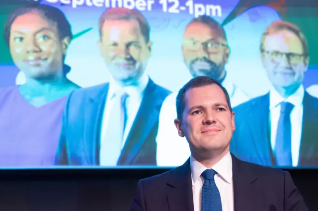 Headshot of Robert Jenrick smiling in front of a screen that has pictures of each MP in the tory leadership race