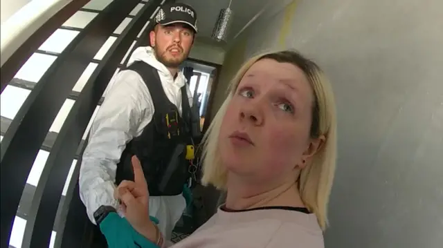 A picture from a body-worn video captured by a police officer. Virginia McCullough is looking over her left shoulder at the officer with a camera. She is wearing a faded pink sweater and being held by a second police officer wearing green gloves, a white forensic suit and black body armour.