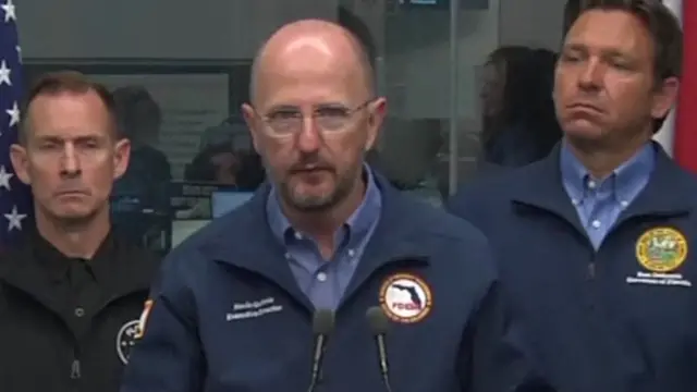 Man in dark blue jacket and shirt speaking behind microphones with two other men stood behind him