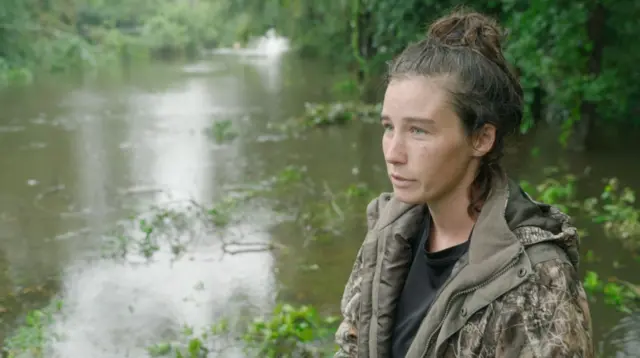 A woman in a green jacket stood in front of a flooded road. She is to the right of the frame and had black hair tied up.