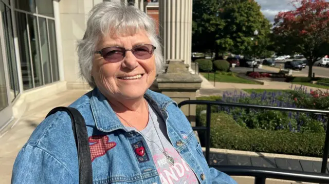 A white older woman stands in the sun and wears a denim jacket