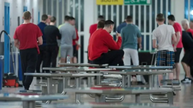 Prisoners in a jail. Some are wearing red jumpers, including one inmate with dark hair who is sitting on a table. All appear to be facing the bars at the far end of the hall.